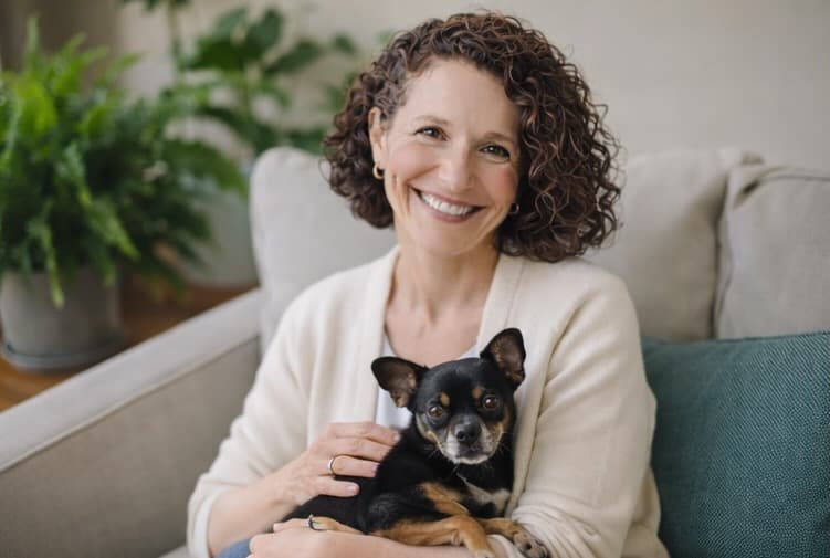 Smiling woman with curly hair sits on a couch holding a black and tan dog.